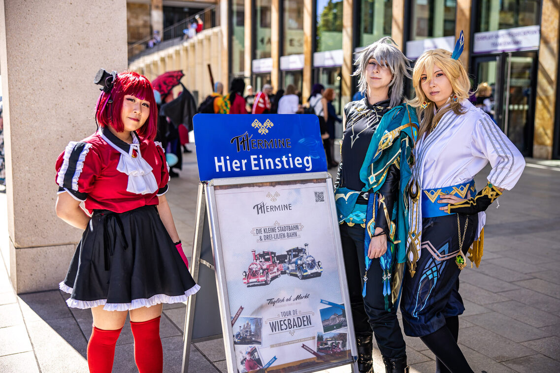 Drei Frauen in Manga-Kostümen stehen vor der THermin Haltestelle