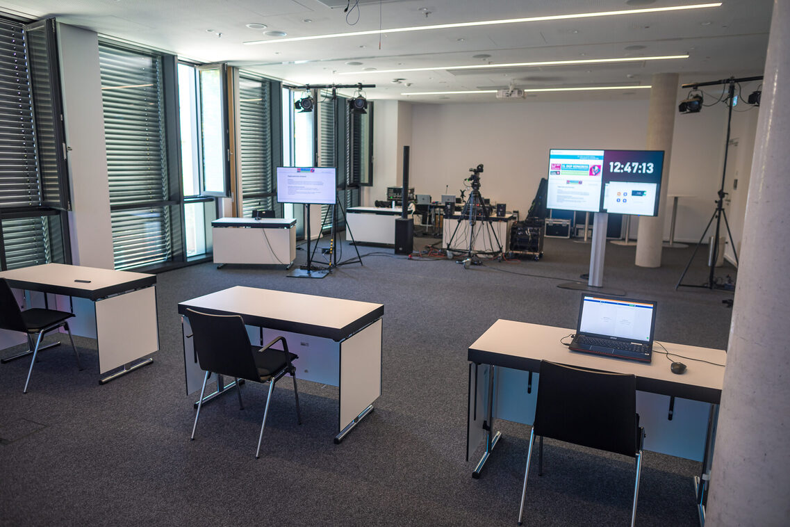 Three individual seats with a table in front of two monitors at a slight distance. Technical equipment behind them.