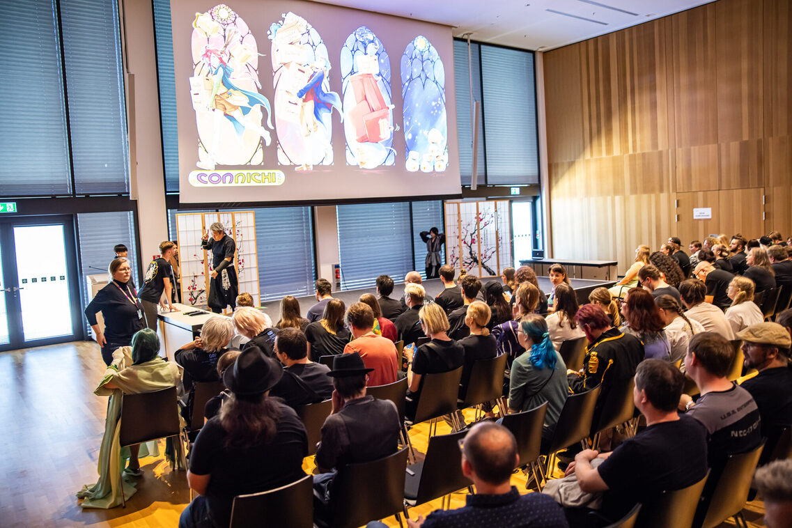 Gäste sitzen in der Reihenbestuhlung und hören den Vortrag zu