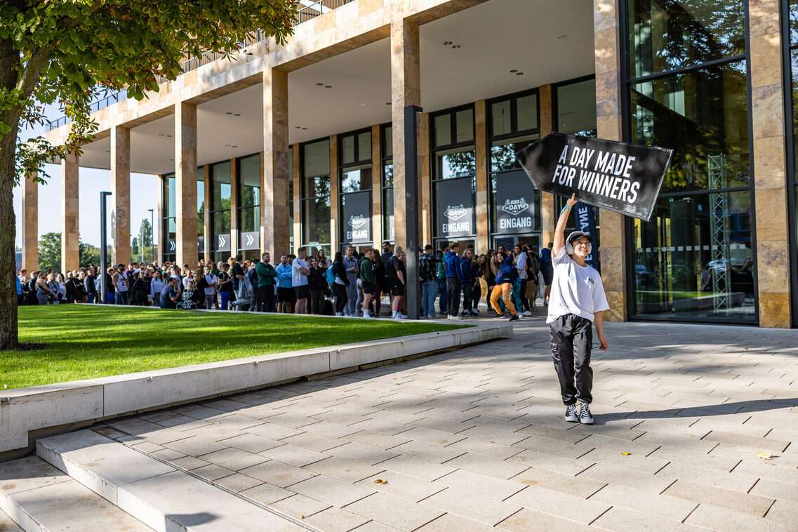 Eine Frau hält ein Schild "A day made for winners" und das Publikum steht in der Warteschlange vor dem RMCC