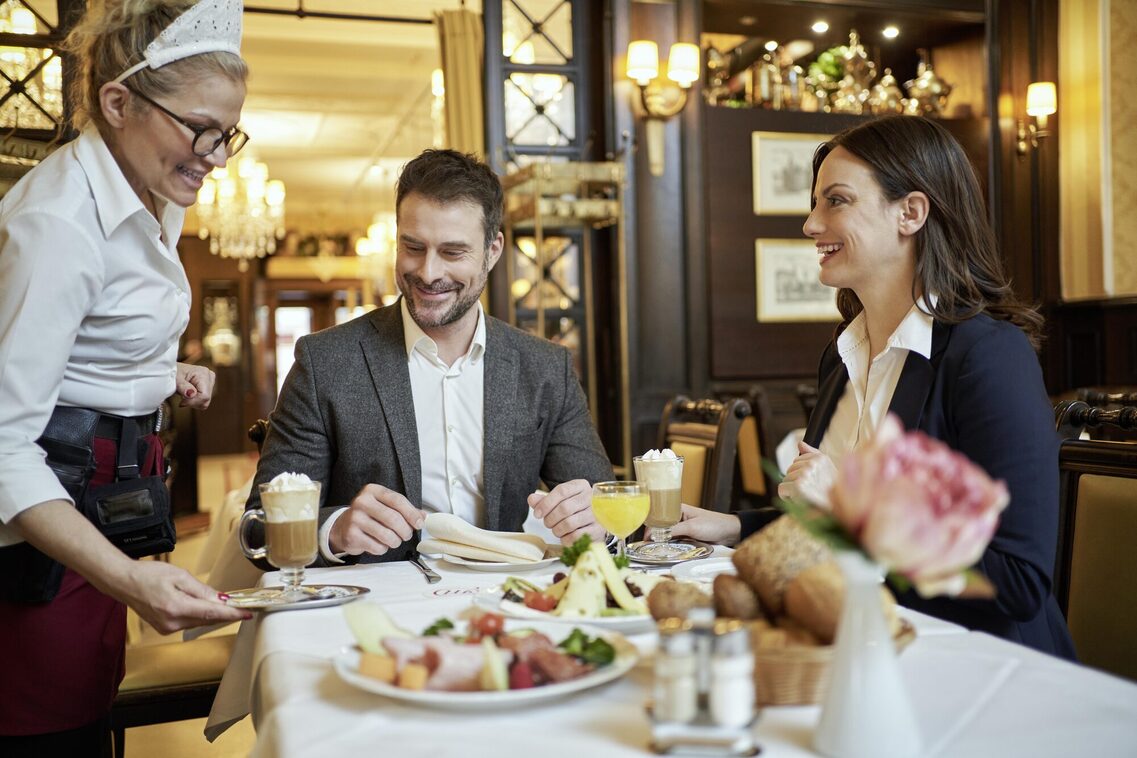Ein Mann und eine Frau sitzen am Tisch und eine Kellnerin bringt Latte Macchiato